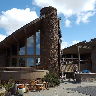 Technician on a tall ladder cleaning high windows beside a stone chimney on a modern home